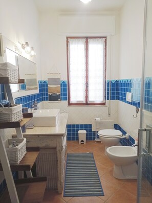 A bathroom with a bathtub, toilet, and sink. There is a window with curtains, a towel rack, and a small stool.