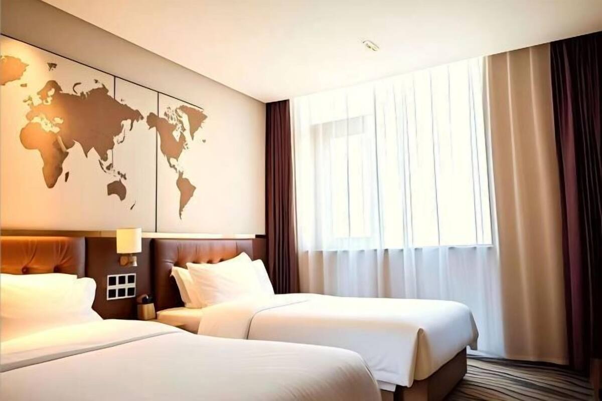 Une chambre d’hôtel avec deux lits, une carte du monde accrochée au mur, une fenêtre avec des rideaux et une tête de lit dotée d’une lampe.