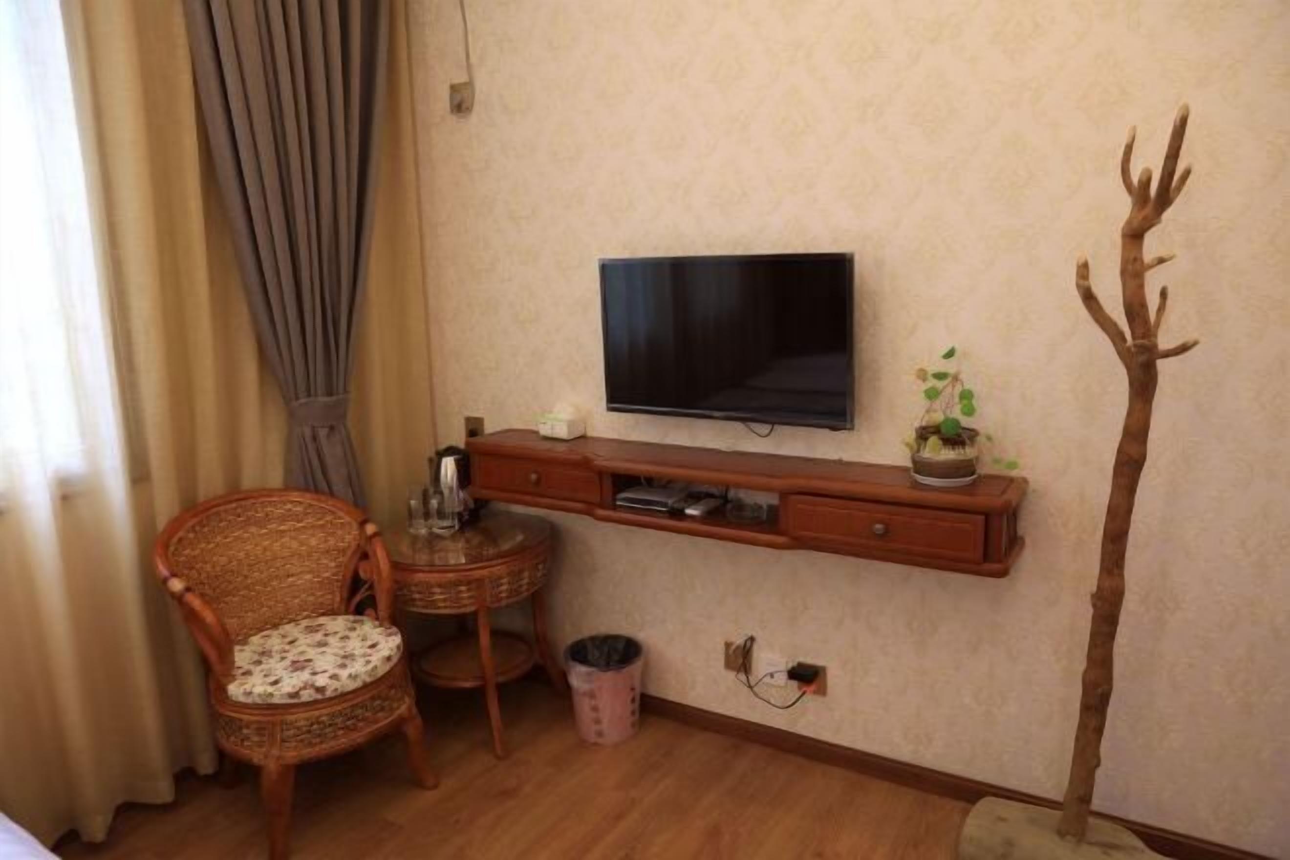 Ein Zimmer mit an der Wand montiertem Fernseher, einem Korbsessel, einem kleinen Tisch mit Pflanze und einem Mülleimer.