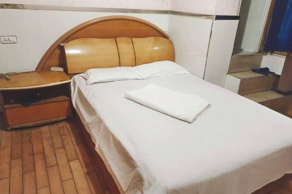 A neatly made bed with a wooden headboard, a wooden nightstand, and a white wall with a power outlet.