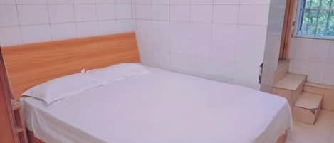 A single bed with a wooden headboard in a room with white tiled walls and a window with a view of greenery.