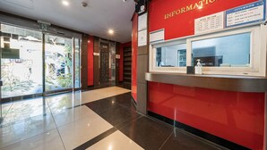 A reception area with a red wall, a glass door leading outside, and a CCTV camera mounted above the reception desk. - Sasang Hotel MU (Busan)