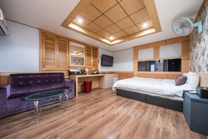 Moderni hotellihuone, jossa on puinen katto, valkoisilla tekstiileillä varustettu sänky, violetti sohva, lasinen sohvapöytä, tietokoneella varustettu työpöytä ja seinälle asennettu televisio.