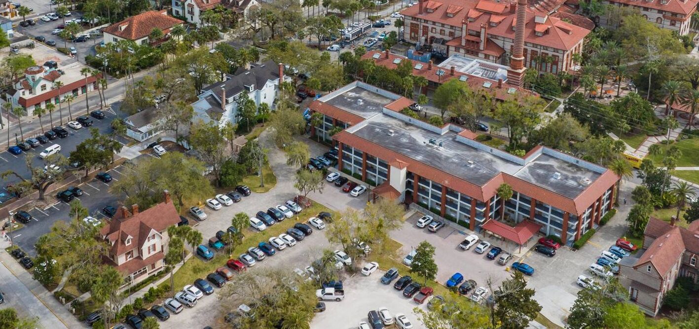An aerial view of a parking lot with multiple rows of parked cars, a large building with a red roof, and surrounding greenery.