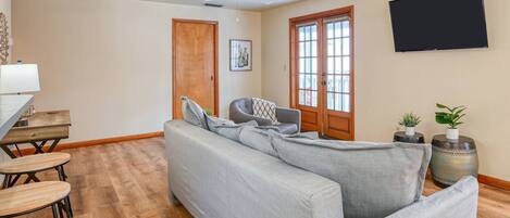A living room with a grey sectional sofa, a wooden dining table with chairs, a ceiling fan, a TV mounted on the wall, and a door with a window.