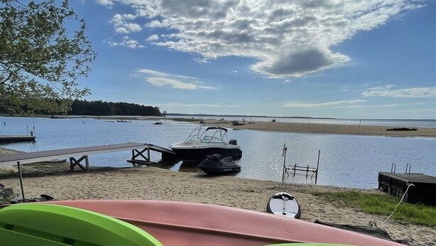A lakeside view with a boat docked near the shore, a wooden dock extending into the water, and a grassy area with a kayak. - Camp Sandbar Limit 12 4 Bedroom Home by RedAwning (Naples)