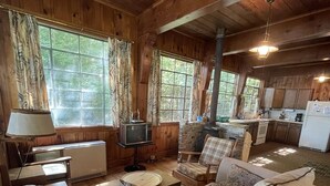 A cozy cabin-style room with a sofa, a small table, a television, and a kitchen area. - Highland Holiday Limit 7-10 3 Bedroom Cottage by RedAwning (Bridgton)