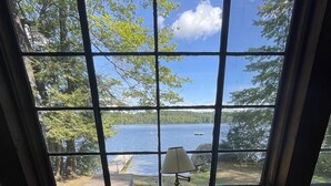 A room with a large window overlooking a lake, a wooden table with books and a lamp, and two chairs. - Highland Holiday Limit 7-10 3 Bedroom Cottage by RedAwning (Bridgton)