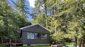 A cabin with a large window, surrounded by trees and a grassy yard. - Highland Holiday Limit 7-10 3 Bedroom Cottage by RedAwning (Bridgton)