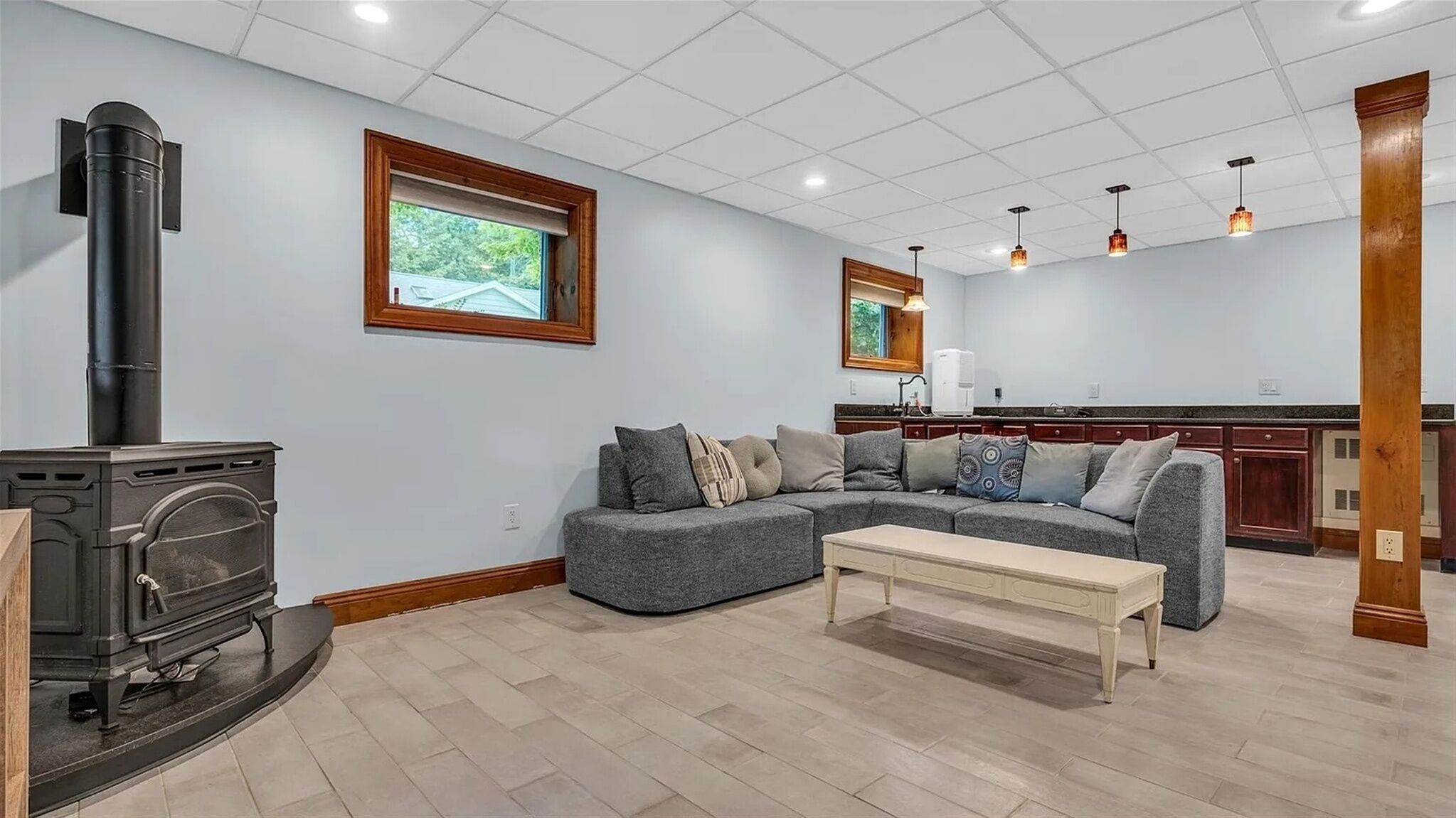 A living room with a fireplace, a sectional sofa, a coffee table, and a kitchen area with wooden cabinets and pendant lights.