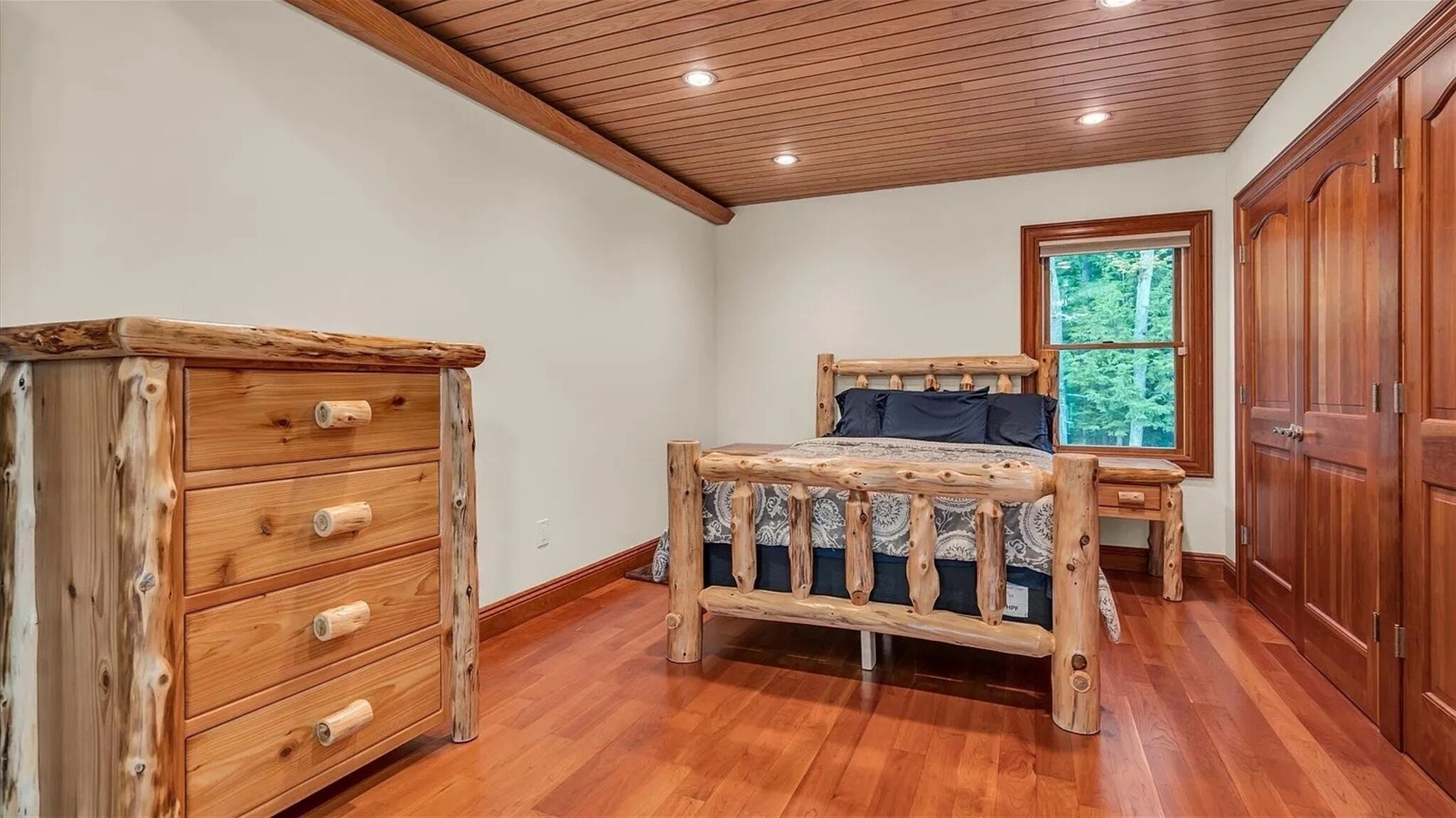 A bedroom with a wooden bed, a dresser, and a window with a view of trees.