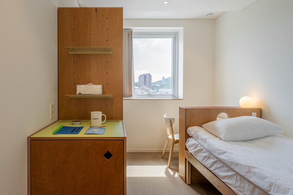 Une chambre d’hôtel compacte, dotée d’un bureau en bois, d’un lit avec une literie blanche, d’une fenêtre donnant sur la ville et d’une petite chaise.