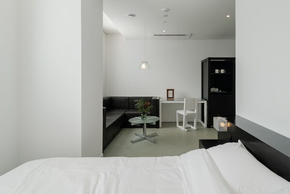 A modern hotel room with a bed, a small round table with a vase, a sofa, a desk, and a chair.