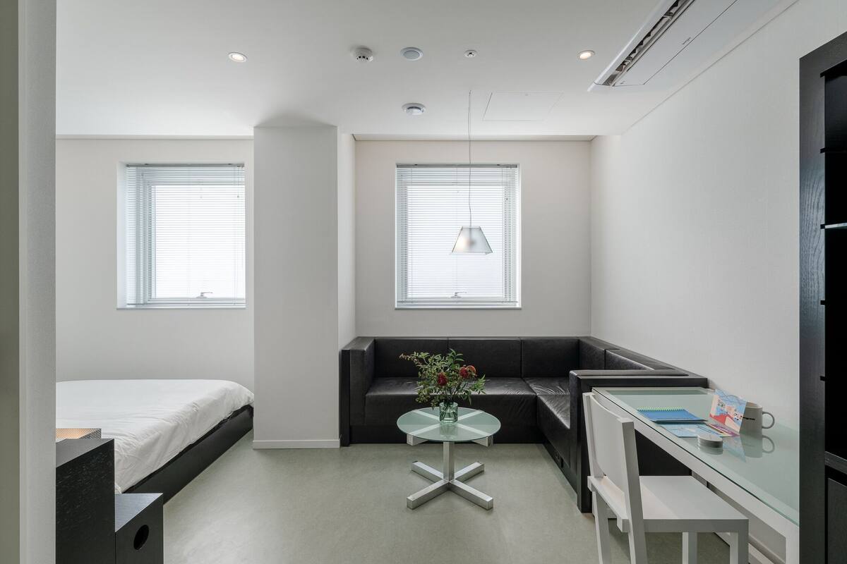 Une chambre moderne et compacte, comprenant un lit, une petite table ronde avec un vase, un bureau avec une chaise et un canapé noir.