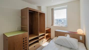 A bedroom with a wooden wardrobe, a bed with white bedding, a bedside table with a lamp, and a window with a city view.