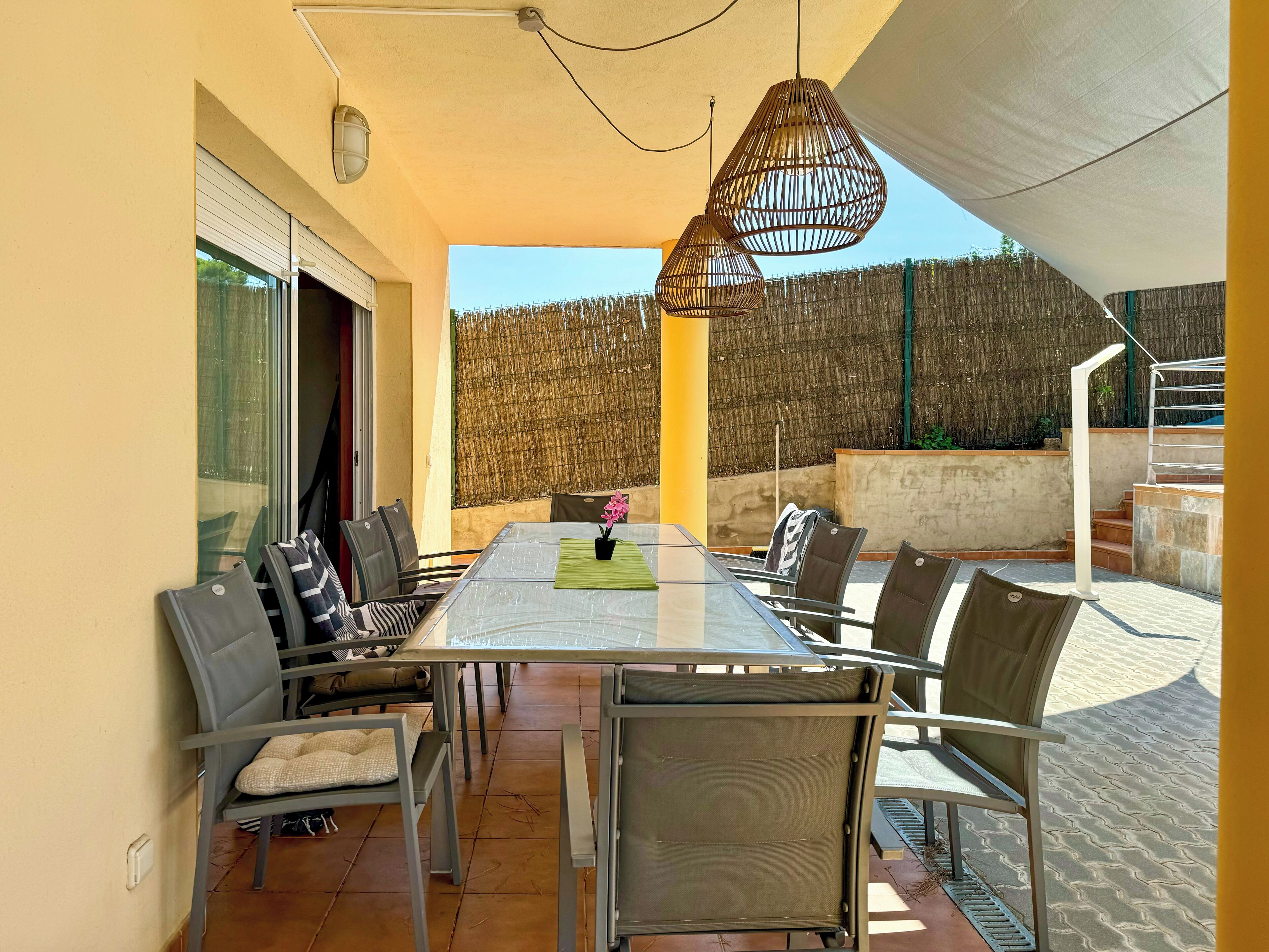 Eine sonnendurchflutete Terrasse mit einem Glastisch, umgeben von Stühlen, einer hängenden Lampe und Blick auf einen Holzzaun.