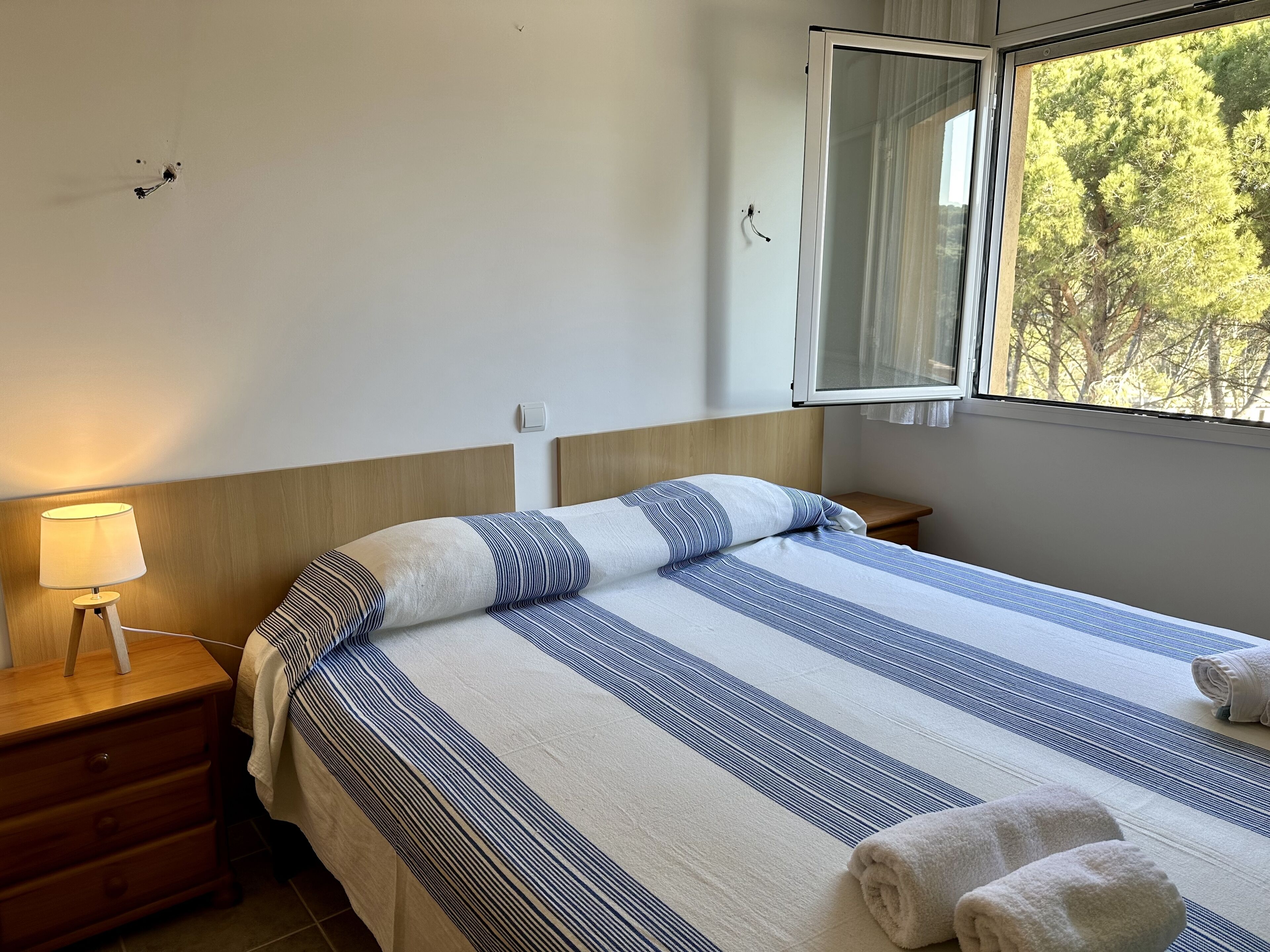 Una cama bien hecha con ropa de cama a rayas, una lámpara de noche, un mesita de noche de madera y una ventana con vistas a árboles.