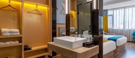 A modern hotel room with a large mirror, a white sink, wooden shelving, and a bed with a blue accent.