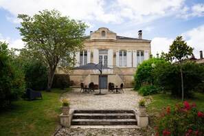 A historic building with a symmetrical facade, a central staircase leading to a covered patio area with outdoor seating, and a well-maintained garden with flowering plants. - Château Papounan (Saint-Estephe)