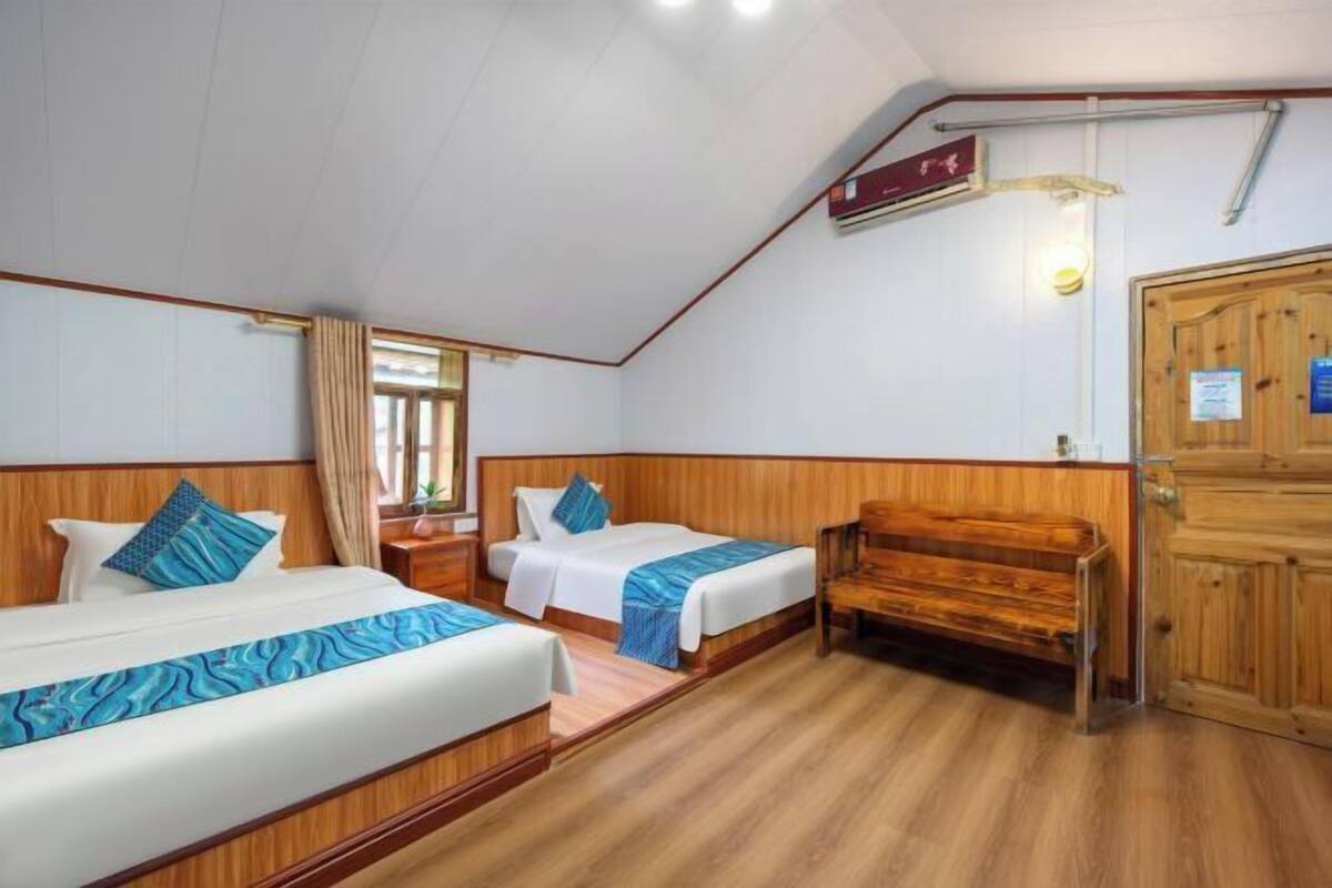 Quarto de hotel com duas camas, soalho em madeira, um banco de madeira, um guarda-roupa e uma janela com cortinas.