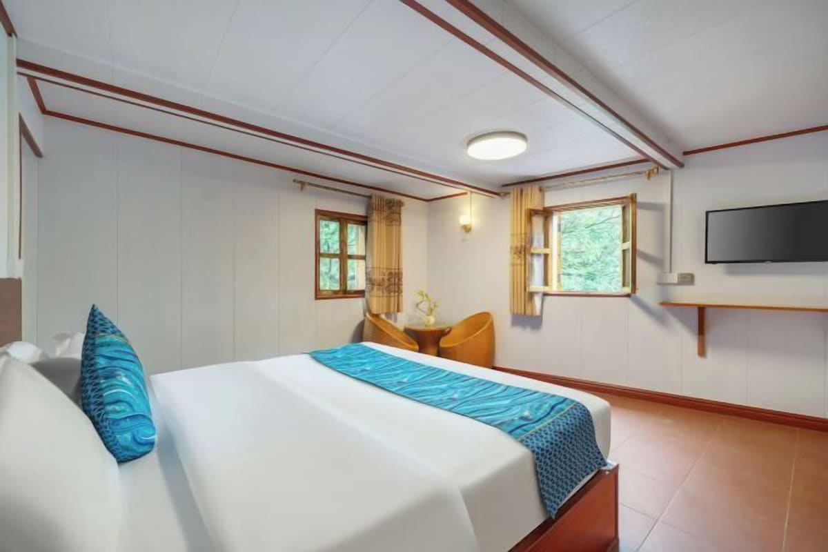 Quarto de hotel com cama, poltrona, televisão e duas janelas.
