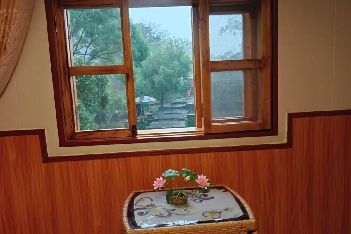 A room with a window overlooking a garden, a small table with a vase of flowers, and wooden paneling on the walls.