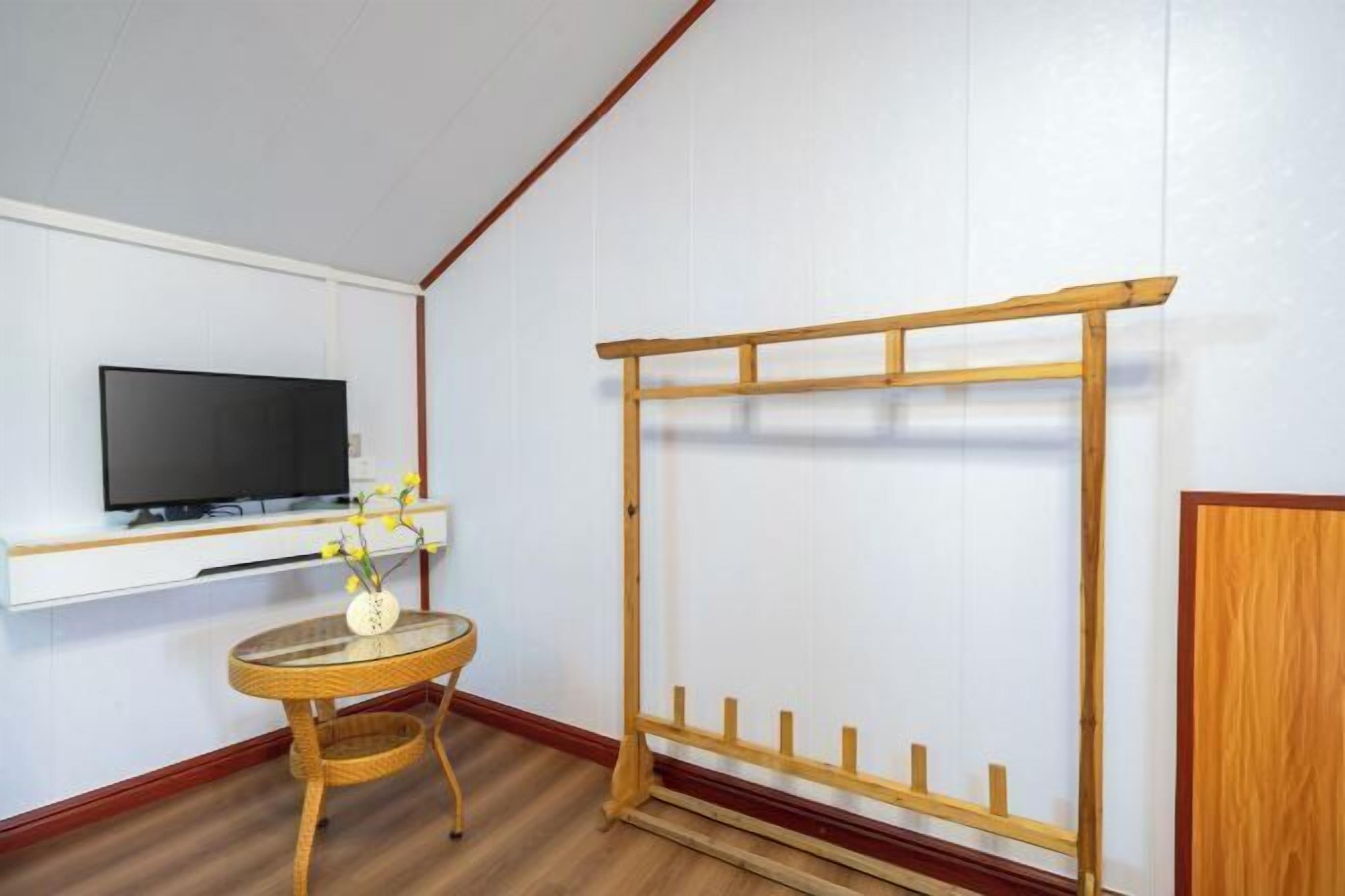 Una habitación con televisión montada en la pared, una pequeña mesa redonda con un jarrón de flores y un perchero de madera.