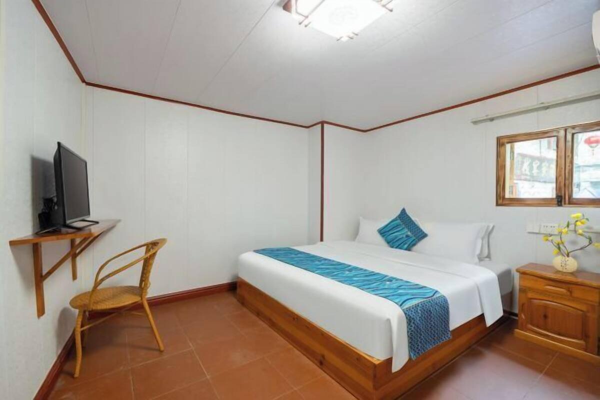 Quarto com cama, escrivaninha com computador, cadeira, janela e mesa de cabeceira com um vaso de flores.