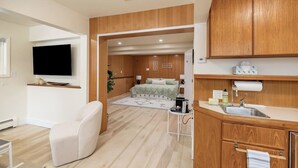 A modern hotel room with a flat-screen TV, a sofa, a bed with a green patterned bedspread, a small table with a lamp, and wooden cabinetry.