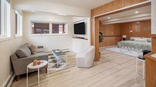 A modern, open-plan living space with a sofa, a coffee table, a TV mounted on the wall, and a bed in the adjacent room.