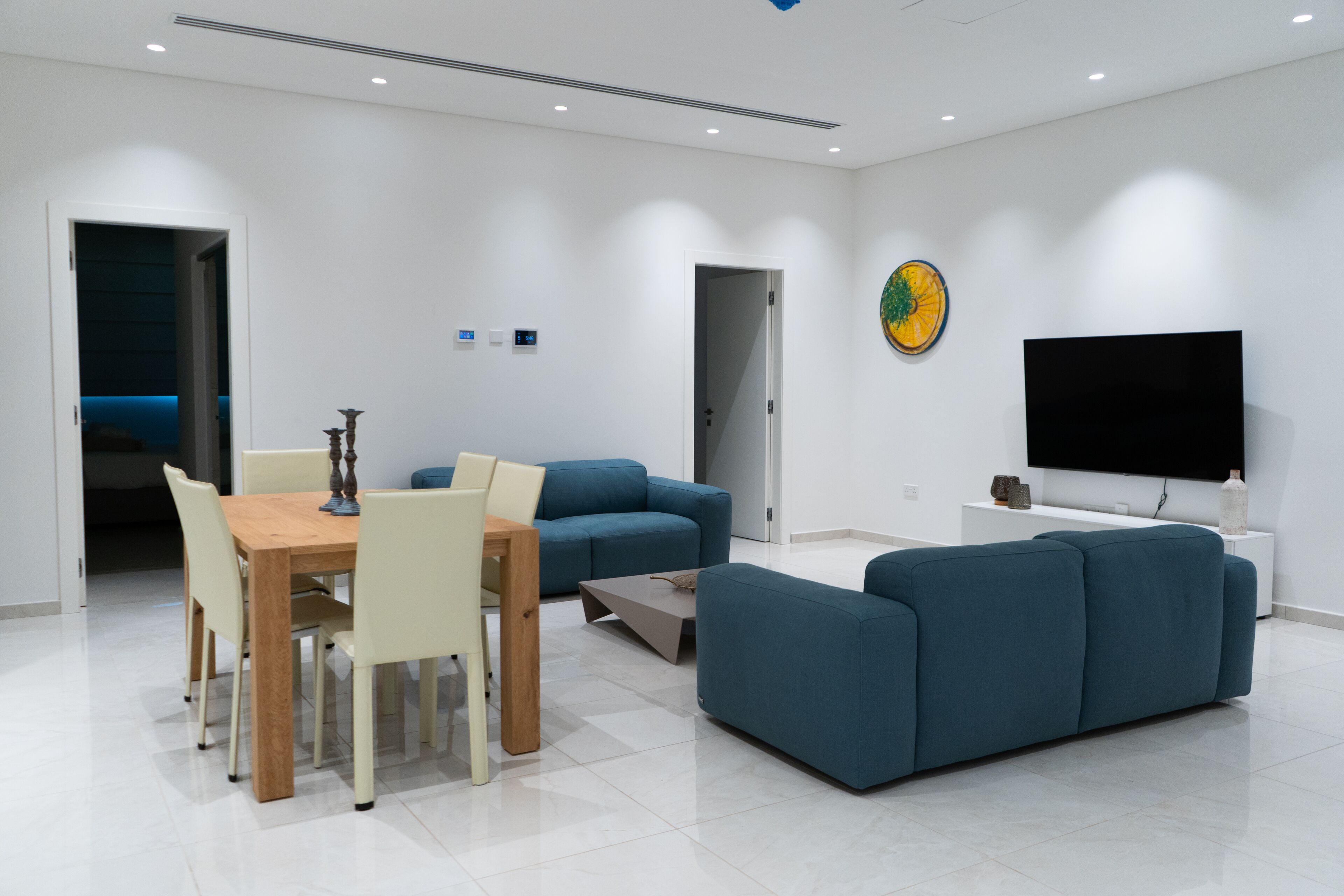 Ein modernes Wohnzimmer mit Essbereich, einem blauen Sofa, einem an der Wand montierten Fernseher und einer Tür zu einem anderen Raum.