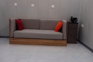 A sofa with two pillows, a coffee machine, and a small cabinet in a room with a tiled floor and a plain wall.
