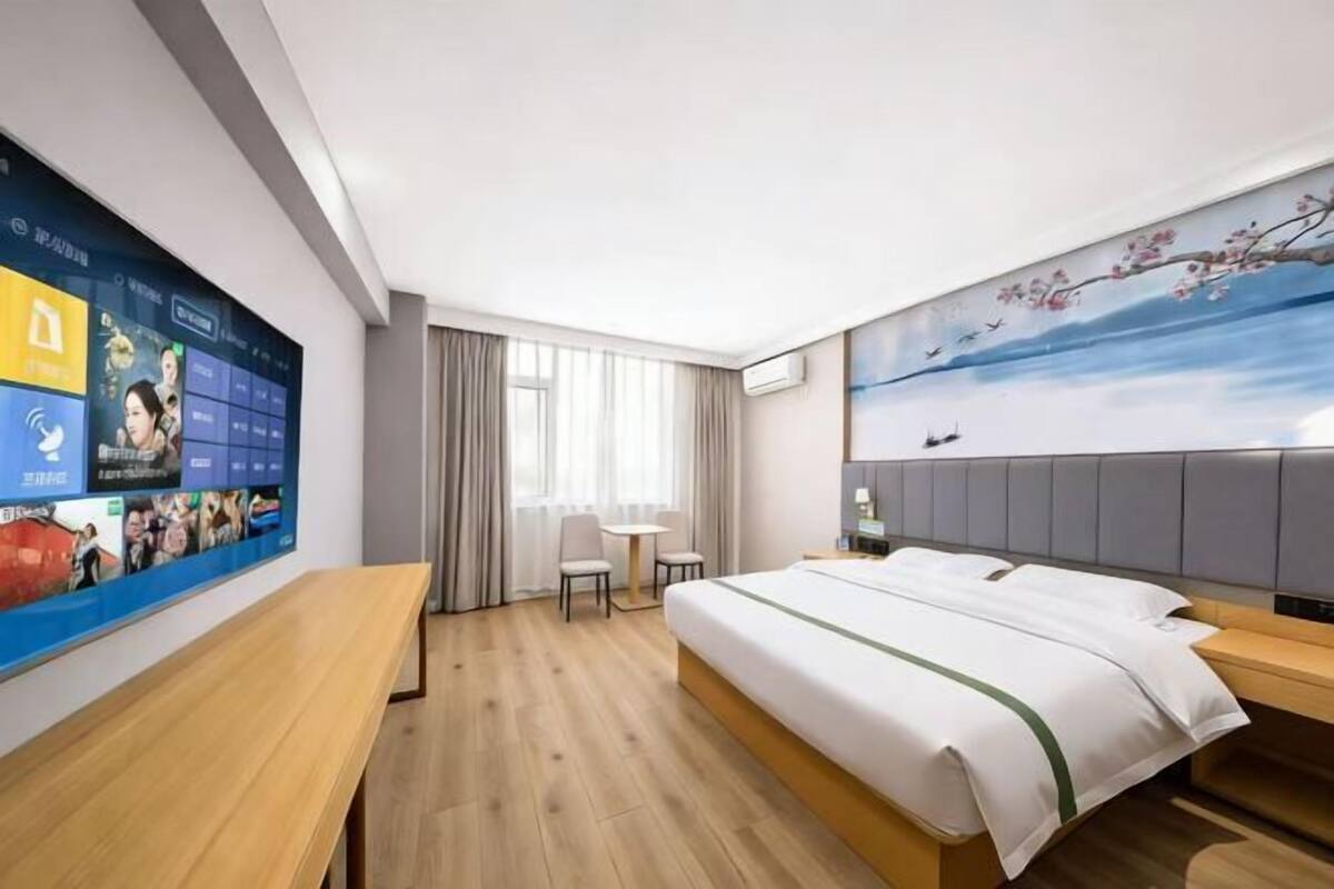Hotellihuone, jossa on suuri digitaalinen näyttö, sänky, työpöytä ja taulu seinällä.