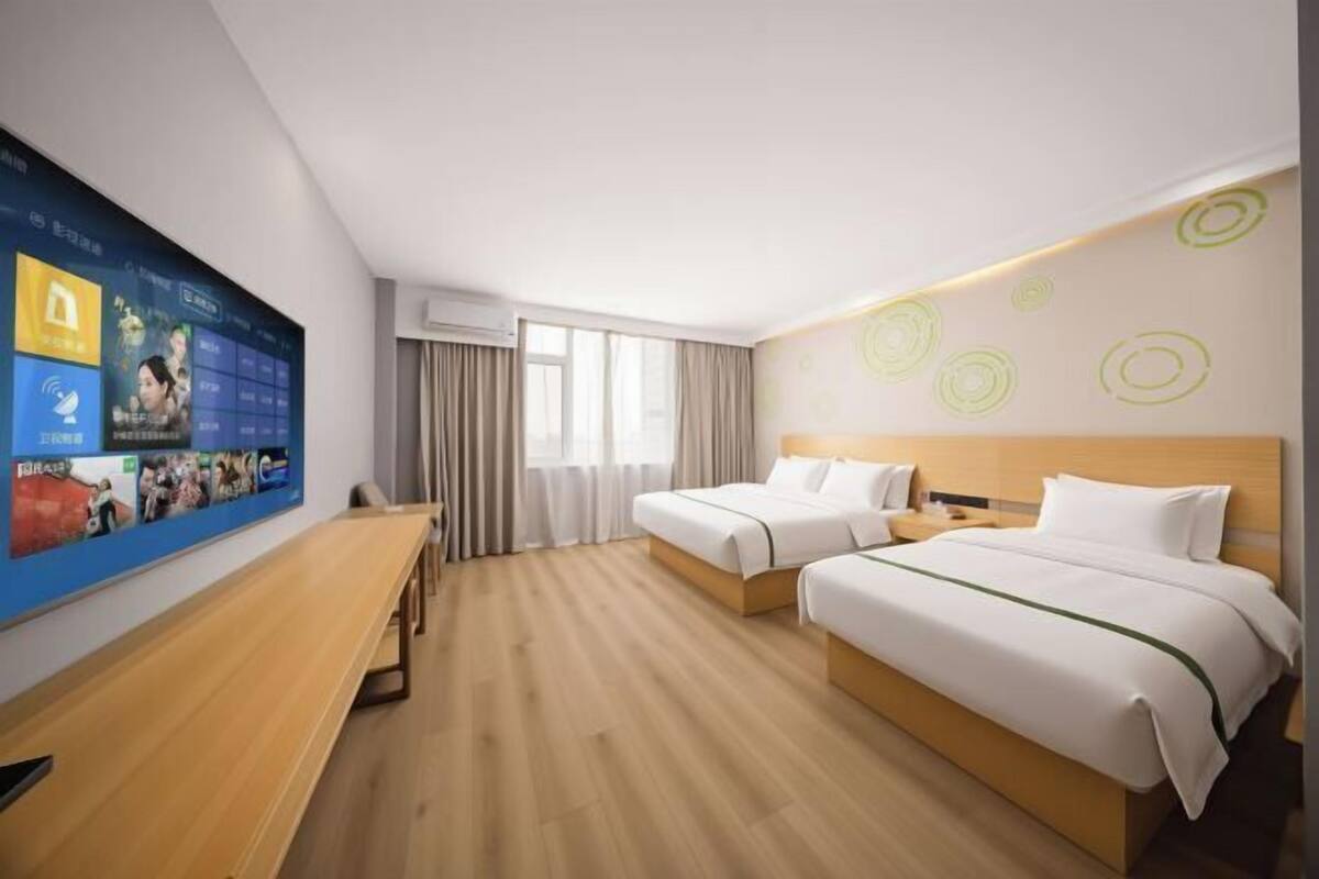 Hotellihuone, jossa on kaksi sänkyä, työpöytä ja suuri digitaalinen näyttö, jolla esitetään sisältöä.