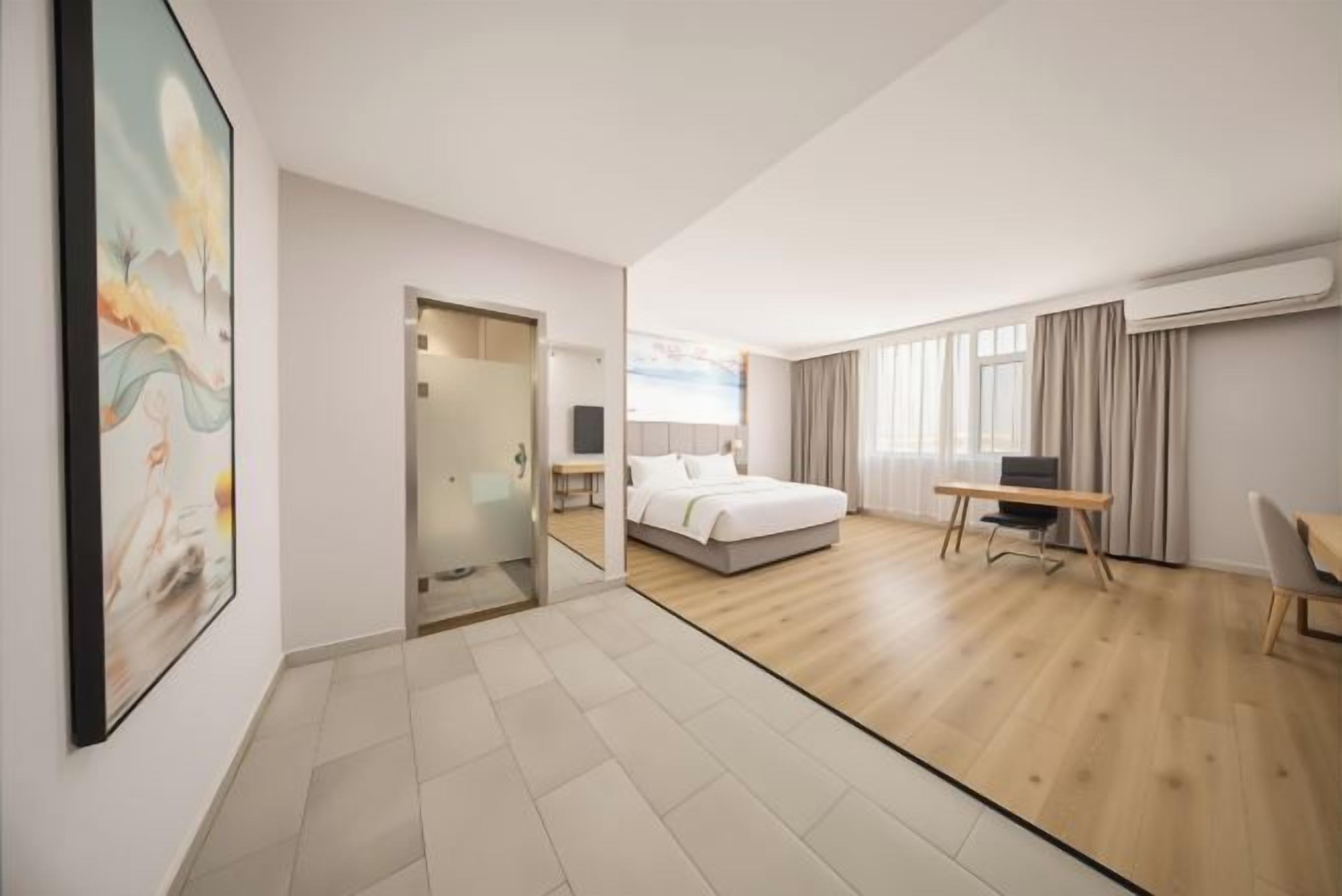 A hotel room with a bed, desk, and chair, a glass door leading to a bathroom, and a large window with curtains.