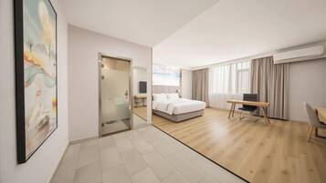 A hotel room with a bed, desk, and chair, a glass door leading to a bathroom, and a large window with curtains.