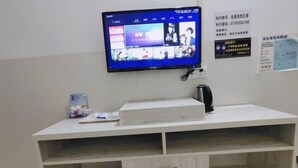 Een kamer met een flatscreen televisie aan de muur, een wit bureau met een lade, en een paar slippers op de vloer.