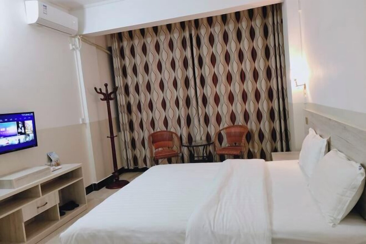A hotel room with a large bed, a TV on a wooden stand, two chairs, and a patterned curtain.