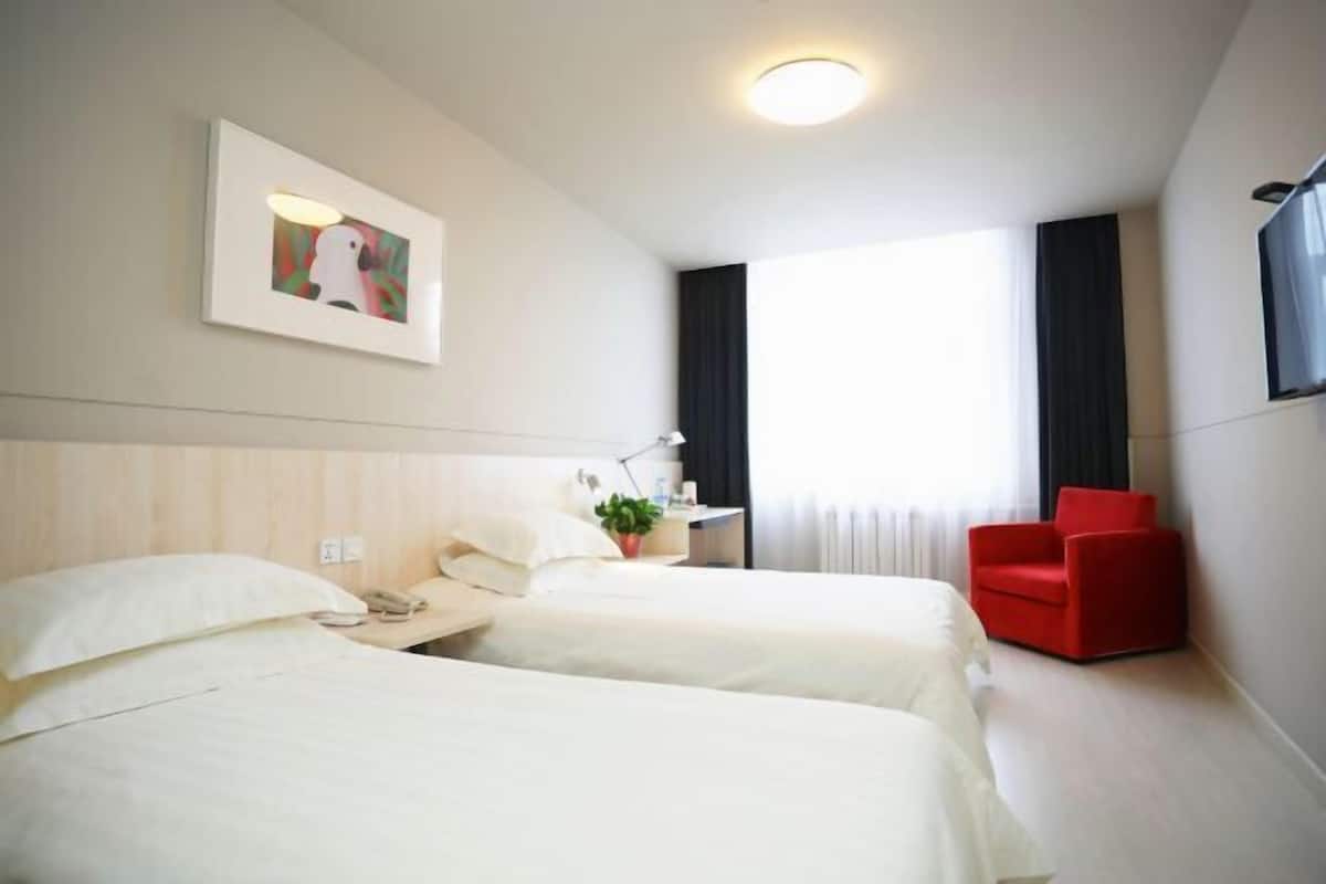 Ein Hotelzimmer mit zwei Betten, einem roten Sessel, einem Schreibtisch mit Lampe, einem Gemälde und einem großen Fenster.
