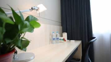 A hotel room with a desk, a lamp, a potted plant, a water bottle, a cup, and a chair.