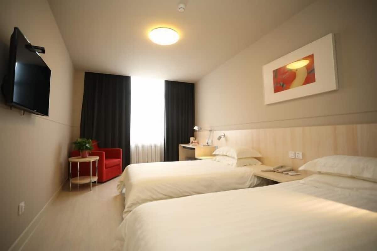 Ein Hotelzimmer mit zwei Betten, einem an der Wand befestigten Fernseher, einem kleinen Tisch mit einer Pflanze, einem roten Sessel und einem Bild an der Wand.