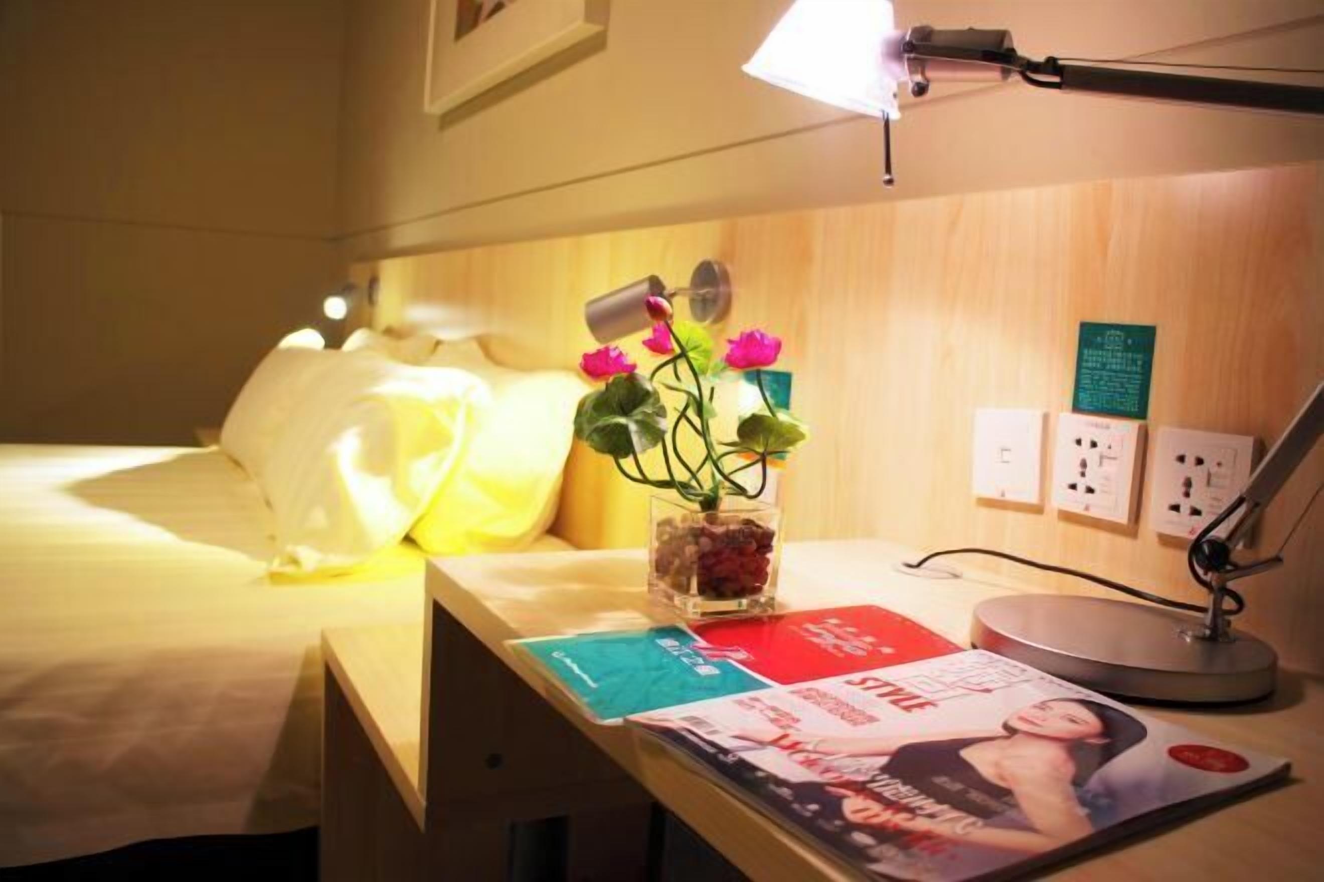 Ein Hotelzimmer mit einem Bett, einem Schreibtisch mit Lampe, einer Vase mit Blumen und einer Zeitschrift.