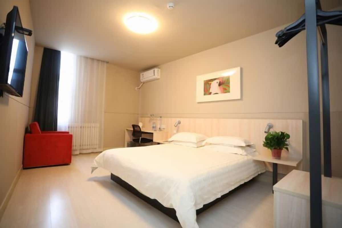 Ein Hotelzimmer mit einem Bett, einem Schreibtisch, einem Stuhl, einem Fernseher, einer Topfpflanze und einem Vorhangfenster.
