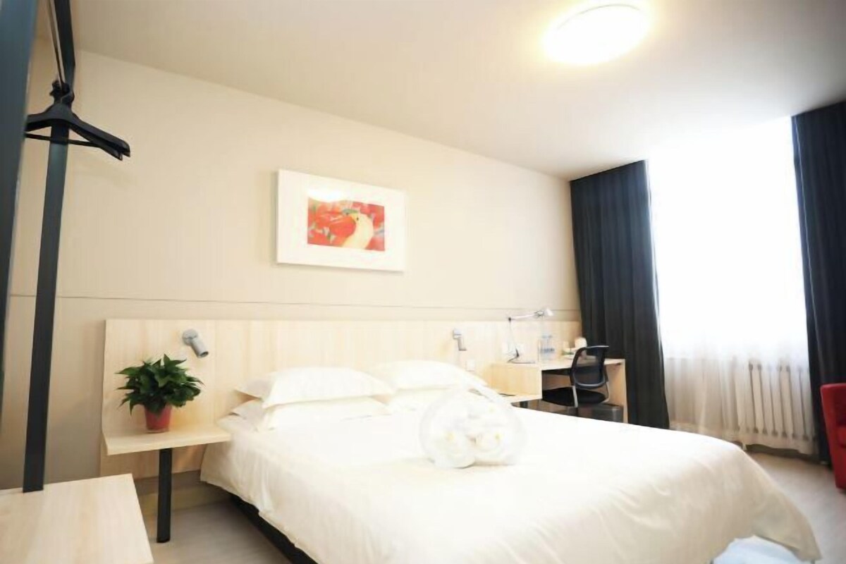 A hotel room with a bed, bedside table, lamp, chair, and a potted plant.