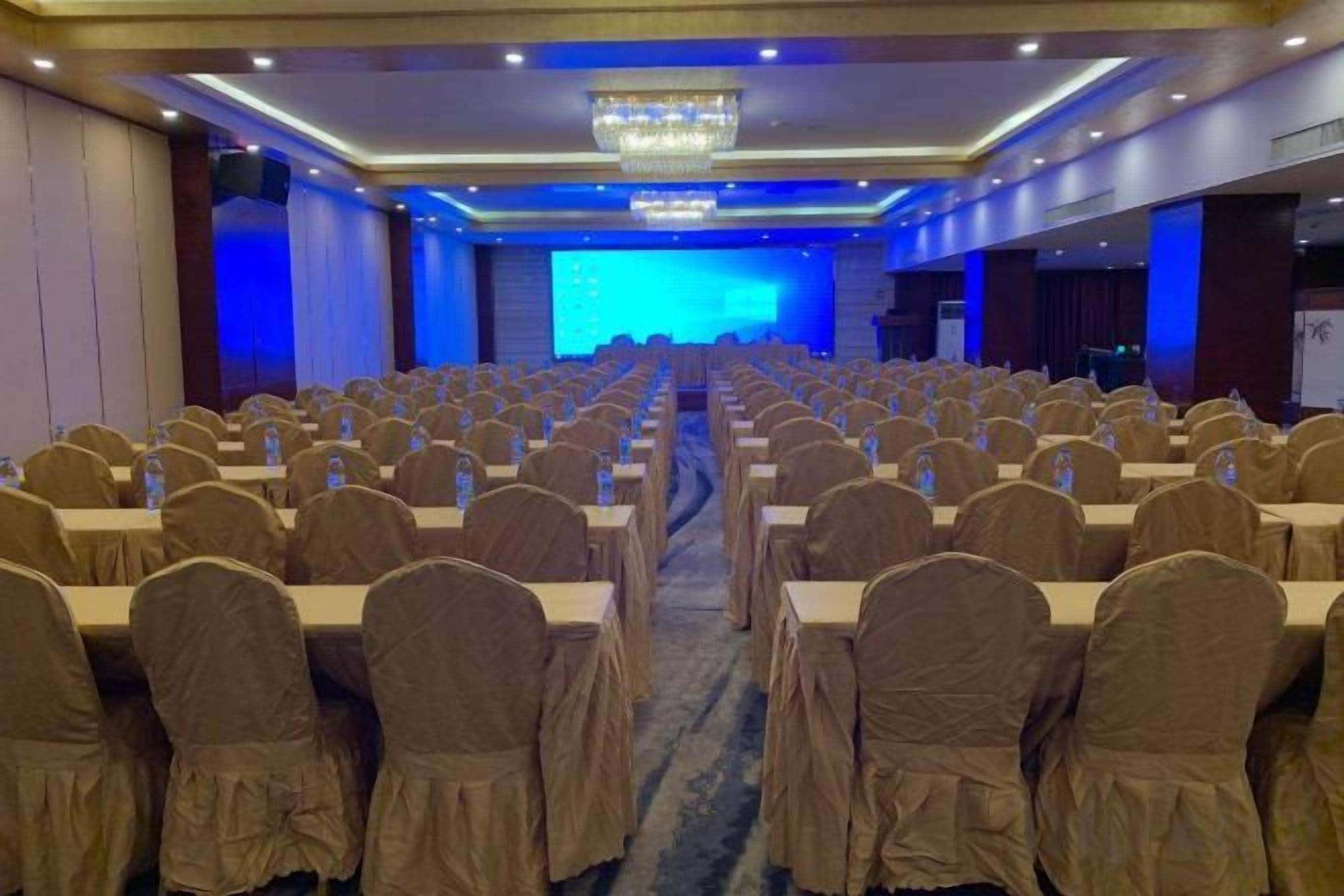 Un salón de conferencias con filas de mesas y sillas, un escenario con pantalla azul y una araña.