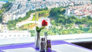 Una habitación con un amplio ventanal que da a un paisaje urbano, un florero con una rosa roja y dos saleros y pimenteros sobre una mesa.