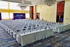 Une salle de conférence avec des rangées de chaises blanches, une longue table et un grand écran.