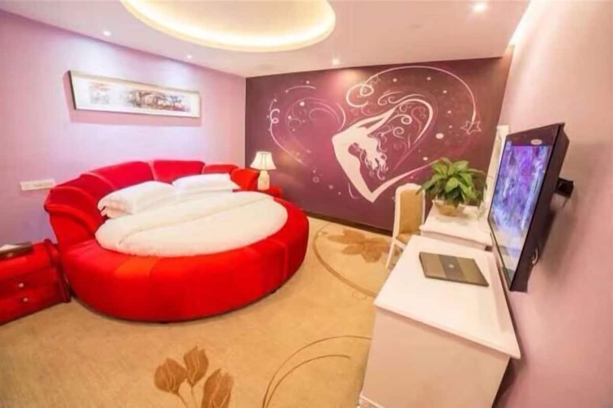 Una habitación con un sofá cama rojo, un televisor de pantalla plana, un escritorio con lámpara y una pared con diseño de corazón.