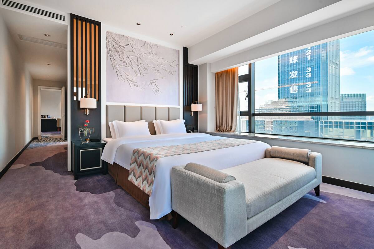 A modern hotel room with a large bed, a sofa, a city view, and a decorative wall.