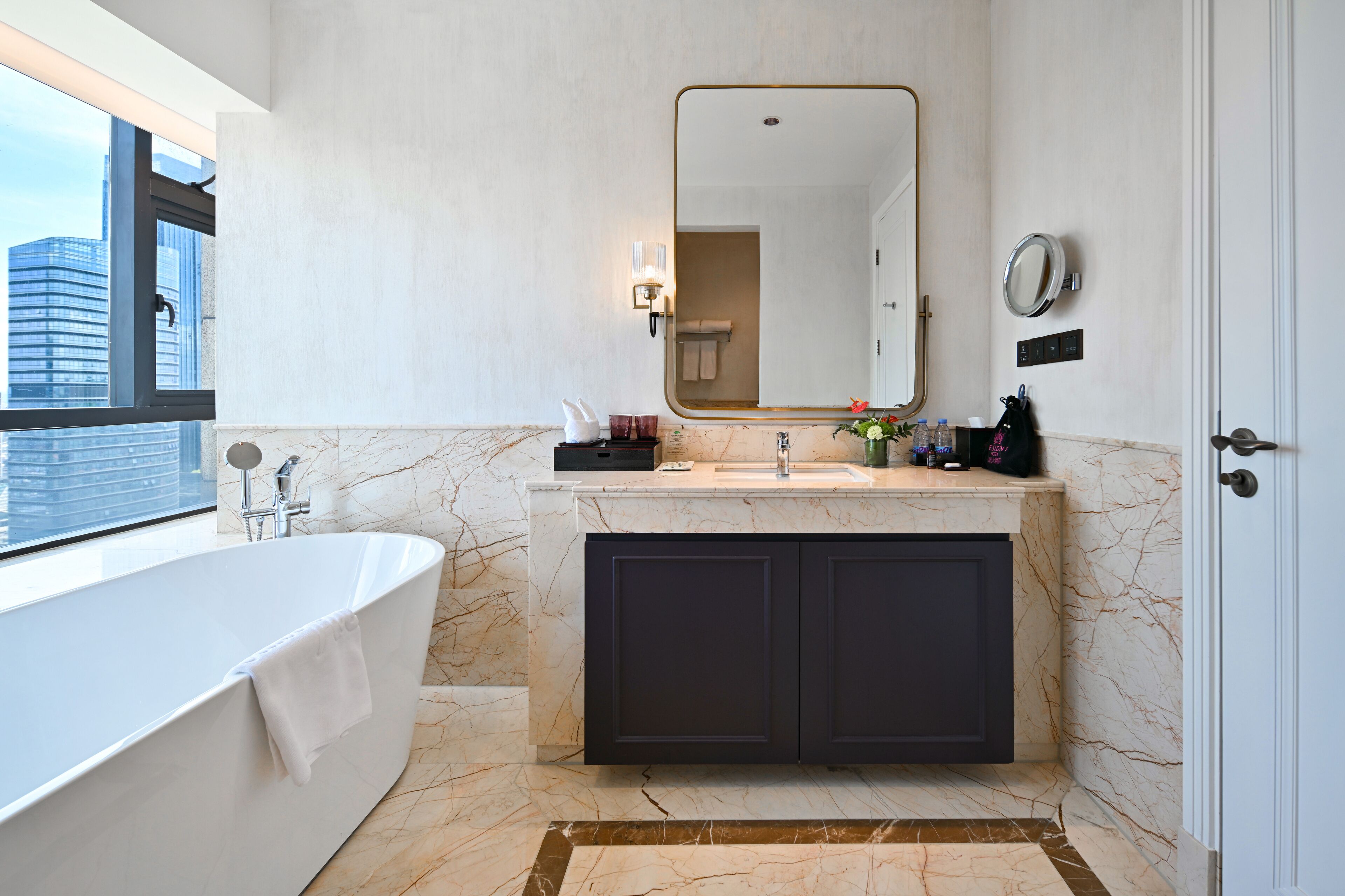 A modern bathroom with a large bathtub, a vanity with dark cabinets, a mirror, and a window offering a city view.